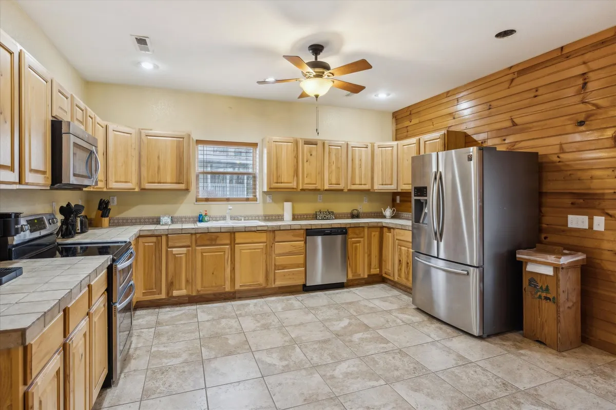 The oversized kitchen is highlighted by wooden cabinets, stainless steel appliances, a central kitchen island, and a ceiling fan overhead. The space feels open and ready for group cooking or entertaining, with plenty of workspace for meal preparation.