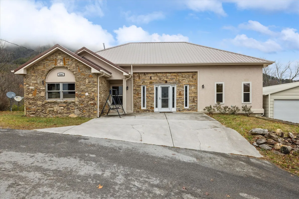 The hero image features the cabin’s striking stone facade and large, light-filled front windows, framed by a symmetrical design. The inviting porch overlooks tranquil mountain scenery, perfect for enjoying stunning sunset views or sipping coffee in the fresh morning air. The setting hints at spacious outdoor areas ideal for family gatherings and relaxing moments.