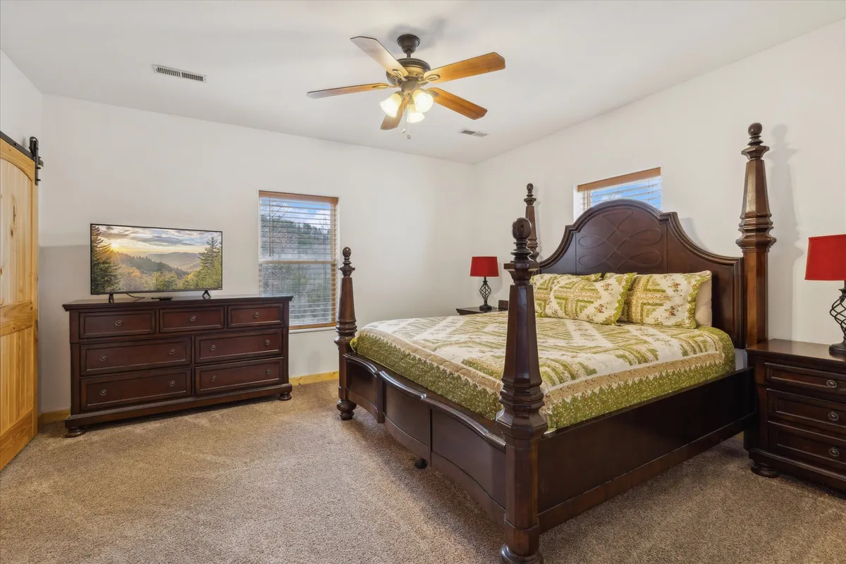 A serene bedroom featuring a king-sized bed with plush bedding, a wooden dresser, nightstands with warm lamps, and a TV on a stand. Gentle natural light filters in, highlighting wood finishes and creating a tranquil, cozy retreat.