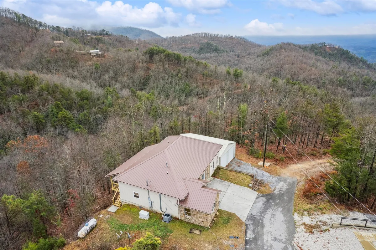 A wide exterior view of the cabin nestled among rolling hills and surrounded by distant mountains. The driveway leads up to the rustic home, and the tranquil setting is enhanced by natural light. The landscape hints at opportunities for outdoor relaxation and scenic mornings.