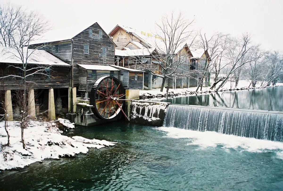 A picturesque scene featuring a rustic water wheel, snow-covered buildings, a flowing waterfall, and a river, all set in a peaceful winter landscape. The setting evokes a sense of discovery and adventure, reminiscent of local attractions like Forbidden Caverns and trout farms.