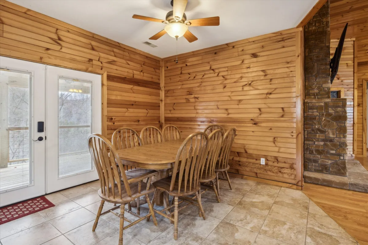 A large wooden dining table surrounded by chairs sits near sliding glass doors, with a stone fireplace in view. The setting is warm and inviting, ideal for group meals and celebrations, accentuated by the glow of the fireplace and a backdrop of wood finishes.
